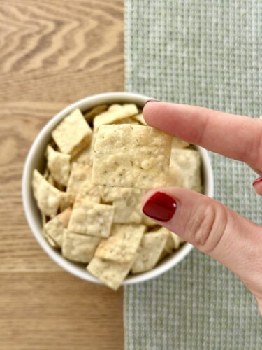 A hand holding a homemade dill cracker above a bowl filled with more crackers.