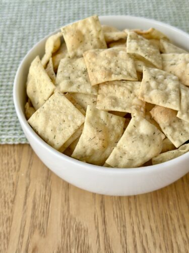 A white bowl filled with homemade dill crackers, arranged in a casual manner, sitting on a wooden table with a green textured cloth in the background.