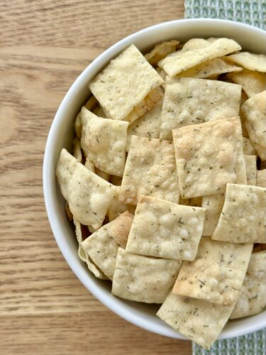 A bowl filled with homemade dill crackers, featuring a mix of golden and lightly browned square-shaped pieces arranged neatly.