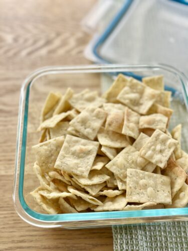 A clear glass container filled with homemade dill crackers, showcasing their crispy texture and irregular shapes, placed on a wooden surface.