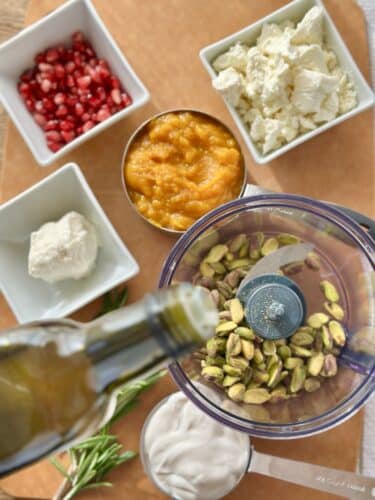 Ingredients for whipped feta pumpkin dip including pumpkin puree, feta cheese, goat cheese, Greek yogurt, pistachios, olive oil, fresh rosemary, and pomegranate seeds arranged on a wooden cutting board.