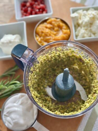 A food processor containing ground pistachios, surrounded by bowls of pomegranate seeds, pumpkin puree, feta cheese, and Greek yogurt on a wooden surface.