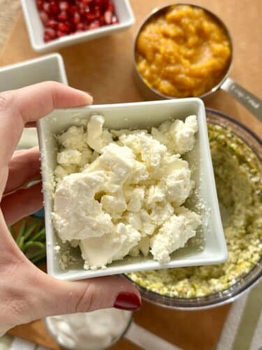 A hand holds a small white bowl filled with crumbled feta cheese, with a food processor containing a blended mixture in the background, along with bowls of pumpkin puree and pomegranate seeds.