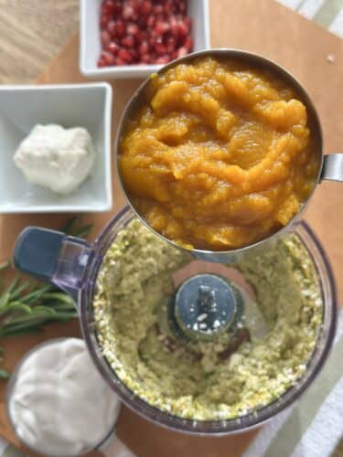 A food processor filled with a creamy mixture being topped with pumpkin puree, alongside bowls of pomegranate seeds and goat cheese, and fresh rosemary, on a wooden surface.