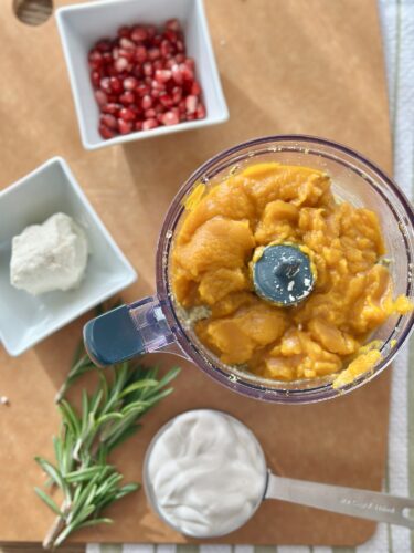 A food processor filled with blended pumpkin puree, surrounded by bowls of goat cheese, Greek yogurt, fresh rosemary, and pomegranate seeds on a wooden surface.