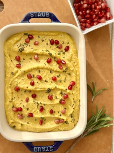 A creamy whipped feta pumpkin dip garnished with pomegranate seeds and fresh rosemary, served in a white baking dish on a wooden surface, with a bowl of additional pomegranate seeds in the background.