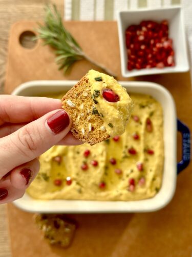 A close-up of a hand holding a crispy cracker topped with creamy whipped feta pumpkin dip, garnished with pomegranate seeds. In the background, a baking dish filled with the dip and a small bowl of pomegranate seeds can be seen, along with a sprig of rosemary.