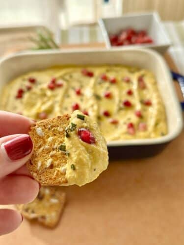 A hand holding a cracker topped with whipped feta pumpkin dip, with pomegranate seeds, in front of a dish of the dip garnished with herbs.