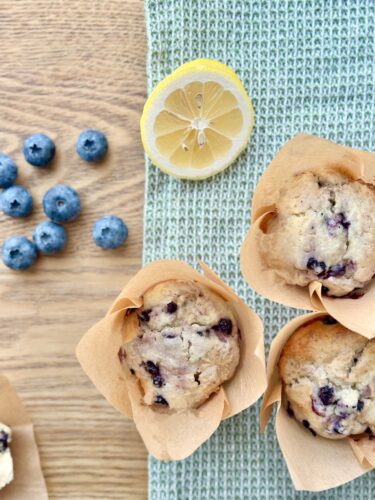Freshly baked lemon blueberry muffins in parchment liners, accompanied by a sliced lemon and scattered blueberries on a textured surface.