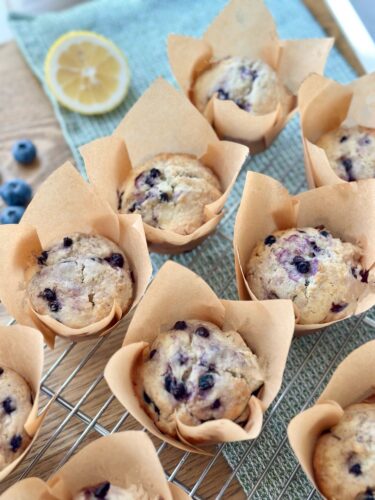 Freshly baked lemon blueberry muffins in parchment paper liners, placed on a cooling rack with blue berries and a lemon slice in the background.
