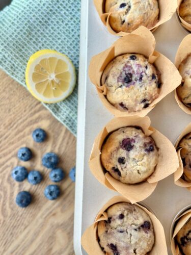 A tray of freshly baked lemon blueberry muffins in brown paper liners, accompanied by a yellow lemon and a few blueberries scattered on a wooden surface.