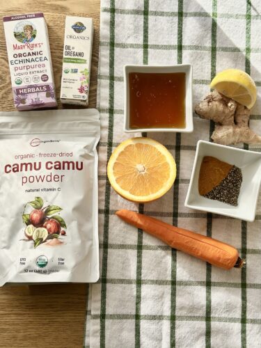 An assortment of ingredients for frozen immunity cubes, including a bag of camu camu powder, fresh ginger, carrot, orange, a bowl of turmeric, and a small container of echinacea liquid extract, all arranged on a checked cloth.