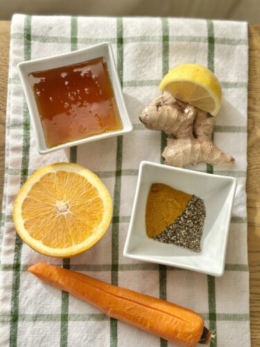 Ingredients for frozen immunity cubes arranged on a checkered cloth: a halved orange, a lemon wedge, a piece of ginger, turmeric, black pepper, honey, and a carrot.