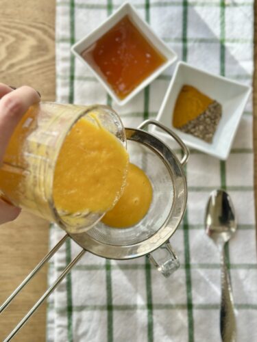 A person pouring a bright orange mixture through a fine mesh strainer into a bowl, with small bowls of honey and spices in the background.