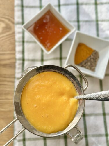 A fine mesh strainer holding a vibrant orange liquid, with bowls of honey, turmeric, and black pepper in the background, resting on a grid-patterned kitchen towel.