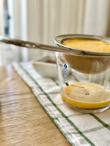 A glass measuring cup with a fine mesh strainer on top, containing a bright orange liquid being poured through the strainer, set on a wooden surface with a green and white checkered cloth.