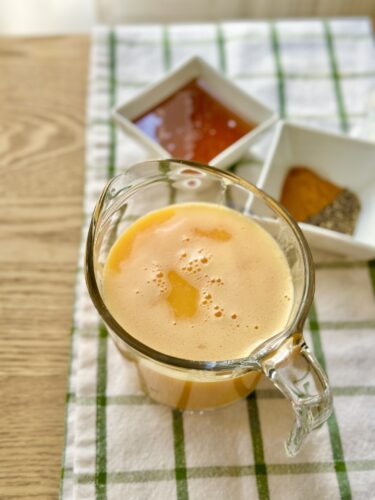A glass pitcher filled with a bright orange liquid, likely a citrus or ginger juice, sits on a wooden table. In the background, small bowls contain honey and spices, hinting at ingredients used in the drink.