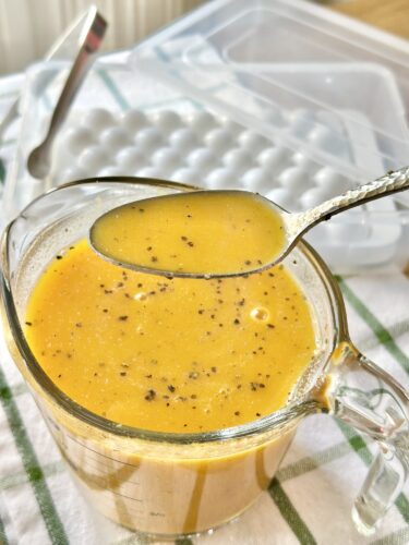 A measuring cup filled with a bright orange liquid that has specks of black pepper on top, with a spoon dipped into it. In the background, a silicone ice tray is visible.