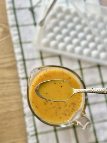 A glass measuring cup filled with a vibrant orange liquid containing spices, alongside a spoon for serving, with a silicone ice cube tray in the background on a checkered cloth.