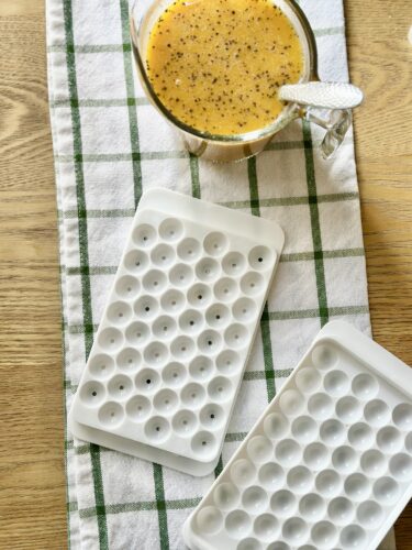 A wooden table with a green and white checkered cloth, featuring a glass of yellow drink topped with black pepper and two ice cube trays beside it.