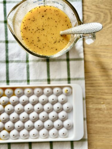 A glass measuring cup filled with a bright orange liquid containing visible black specks, resting on a checkered towel, with a silicone ice cube tray filled with rounded mold sections in the background.