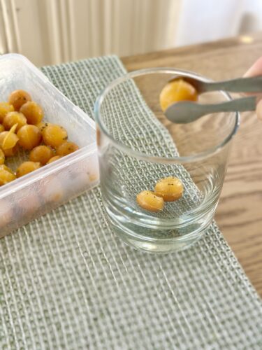A person using tweezers to pick up frozen immunity cubes from a container, placing them into a glass on a textured table surface.