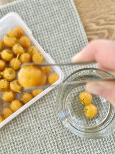 A hand using a fork to pick up yellow balls from a clear container, with a glass jar set on a textured surface.