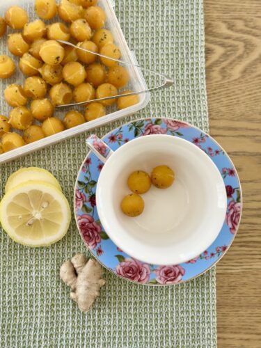 A decorative plate with a floral design holds three yellow immunity cubes beside a halved lemon and fresh ginger on a textured green tablecloth. A container of yellow cubes is placed in the background.
