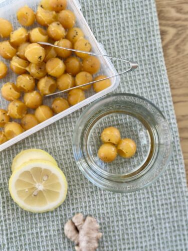 A small container of orange immunity cubes sits next to a glass of water, with lemon slices and fresh ginger nearby on a textured surface.