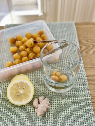 A glass bowl with frozen citrus-ginger immunity cubes next to a halved lemon and a piece of fresh ginger, set on a textured green placemat.