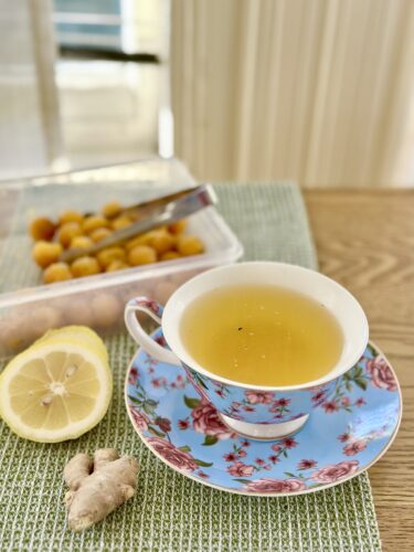 A floral teacup on a wooden table holds a warm drink, surrounded by a lemon half, fresh ginger, and a container of frozen immunity cubes.