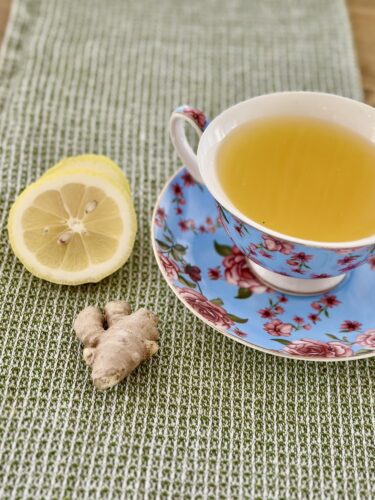 A cup of tea with lemon slices and fresh ginger on a floral-patterned saucer, placed on a textured green placemat.