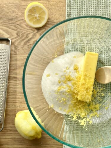 A mixing bowl containing sugar, a stick of butter, and lemon zest, with a halved lemon and a zester nearby.