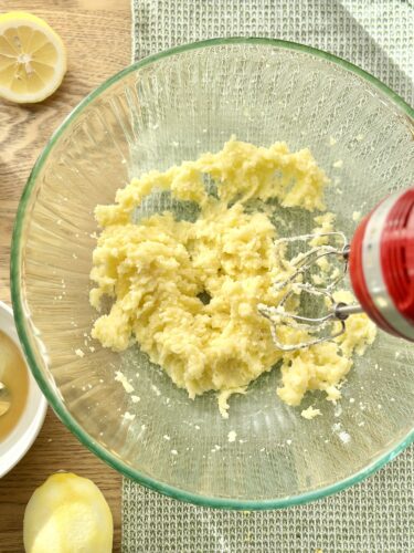 A mixing bowl containing creamed butter and sugar with a hand mixer, surrounded by halved lemons.