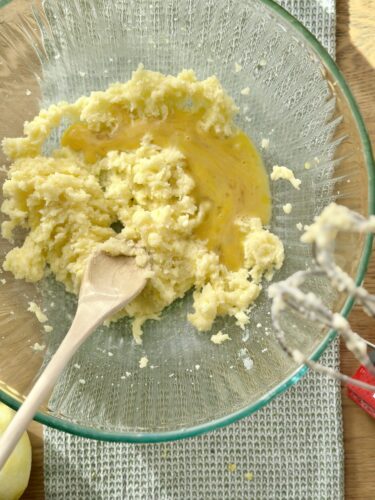 A glass mixing bowl containing a mixture of creamed butter and sugar with an egg added, beside a wooden spoon and mixer attachments.