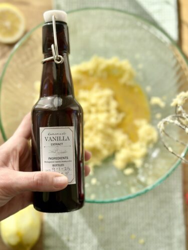 A hand holding a bottle of vanilla extract in front of a mixing bowl with creamed butter and lemon zest, showcasing the ingredients for lemon blueberry muffins.