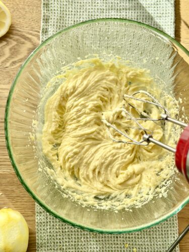 A glass mixing bowl containing creamy, whipped butter and sugar, with a hand mixer resting against the bowl. Lemons and a kitchen towel are visible in the background.