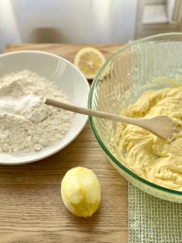 A wooden spoon resting in a bowl of yellow batter, with a plate of flour and a whole lemon in the background, set on a wooden surface.