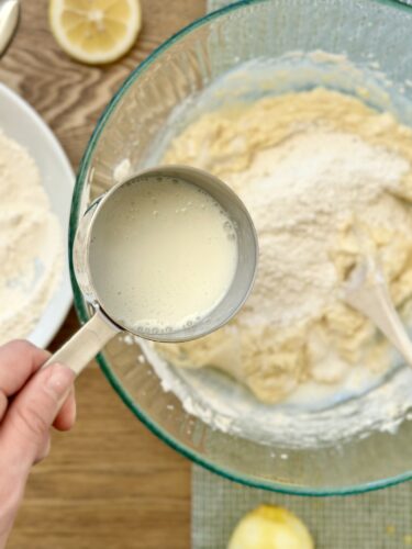 A hand holding a measuring cup filled with buttermilk, poised above a mixing bowl containing muffin batter, with scattered flour and lemon halves in the background.