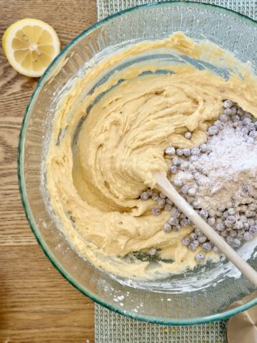 A mixing bowl containing thick batter for lemon blueberry muffins, with flour-coated blueberries being added in. A sliced lemon is visible in the background.