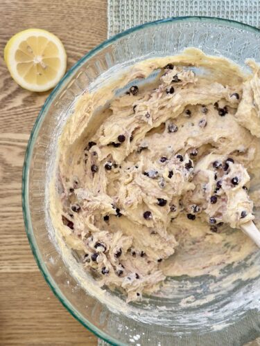 A glass bowl filled with thick lemon blueberry muffin batter, studded with frozen blueberries, next to a halved lemon on a wooden surface.