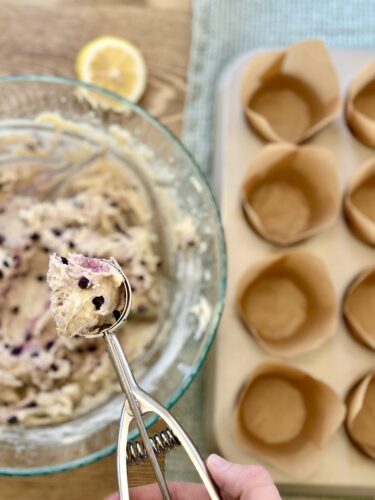 A hand holding a cookie scoop filled with lemon blueberry muffin batter above a bowl of the batter, with lemon slices and empty parchment-lined muffin cups in the background.