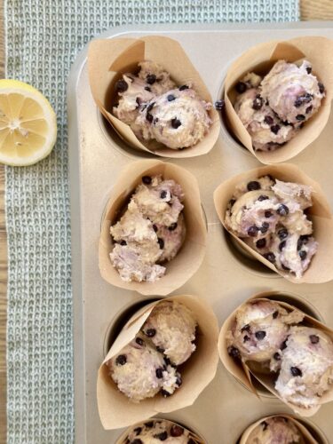 A muffin tin filled with unbaked lemon blueberry muffin batter, in parchment liners, alongside a lemon half on a textured green surface.