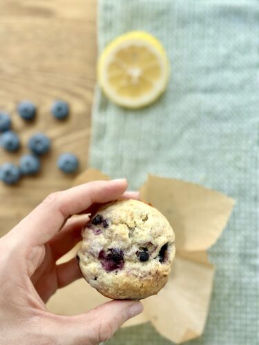 A person holding a lemon blueberry muffin in front of a slice of lemon and scattered blueberries on a wooden surface.