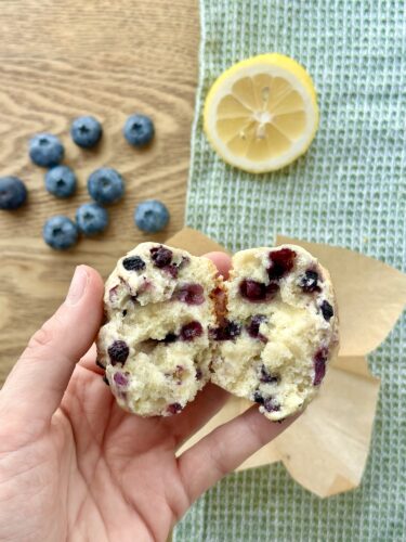 A hand holding a split lemon blueberry muffin, revealing its soft texture and blueberry filling, with scattered blueberries and a lemon slice in the background.