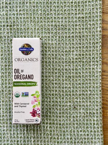 A box of oil of oregano liquid herbal supplement with a white and green design, placed on a textured green woven fabric background.