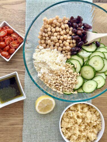 A large glass bowl filled with ingredients for Greek couscous salad, including sliced cucumbers, crumbled feta, chickpeas, Kalamata olives, and pine nuts, with a small dish of slow-roasted cherry tomatoes and a lemon half nearby.