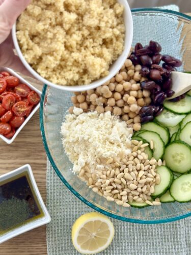 A bowl filled with ingredients for Greek couscous salad, including cooked couscous, chickpeas, Kalamata olives, crumbled feta cheese, sliced cucumbers, and pine nuts, with cherry tomatoes and dressing on the side.