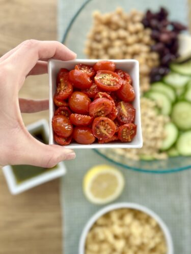 A hand holding a bowl of sliced slow-roasted cherry tomatoes with a background of prepared ingredients for Greek couscous salad, including cucumbers, chickpeas, Kalamata olives, and a lemon.