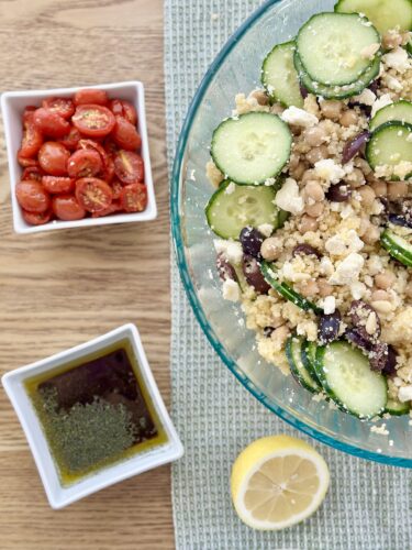 A glass bowl filled with Greek couscous salad featuring sliced cucumbers, chickpeas, crumbled feta cheese, and Kalamata olives, accompanied by bowls of slow-roasted cherry tomatoes and dressing, with a lemon half placed nearby.